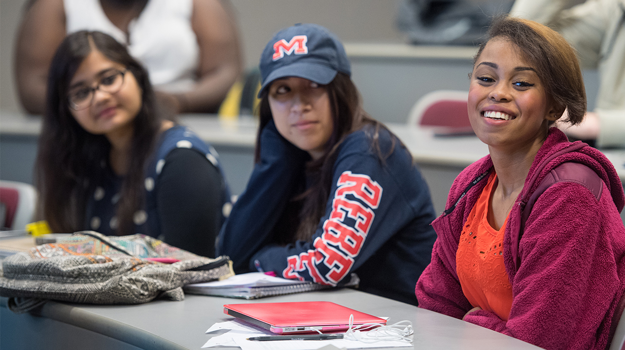 Students in a classroom. One focused student is smiling.