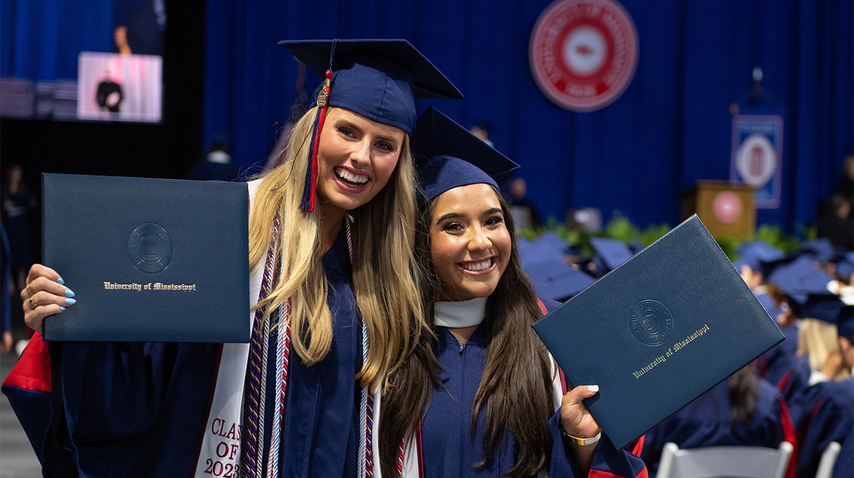 Two female students during the undergraduate commencement holding up their degrees.