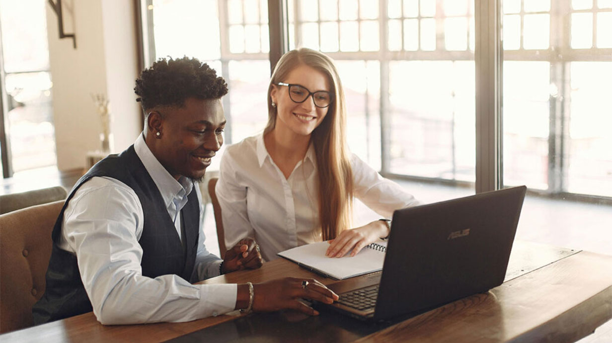 A male and female sitting next to each other looking at a laptop on a desk. The image depicts mentorship.