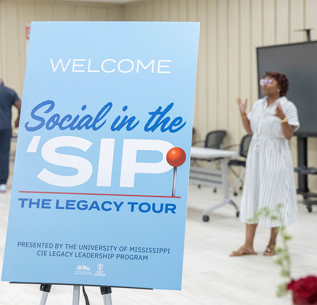 A standee with text reads, "Welcome. Social in the 'Sip Legacy Tour. Presented by the University of Mississippi CIE Legacy Leadership Program." Angelica Owens is in the background giving a speech.