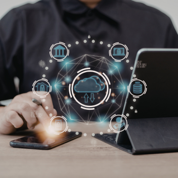 Canva Image: A man is seated at a desk, using a smartphone and a laptop. Surrounding him is a digital interface displaying various AI-related icons and symbols, indicating a focus on artificial intelligence and technology. The setting appears to be a modern workspace.