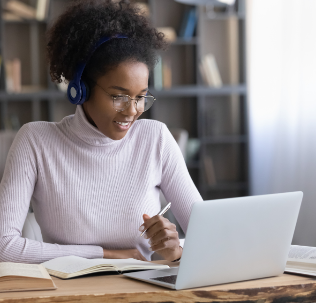 A female student wearing headphones while taking notes in front of a laptop.