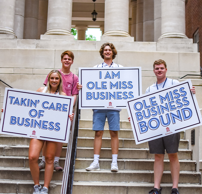 A group of four students outside Holman Hall holding up signs saying: Takin' care of business, I am Ole Miss Business, and Ole Miss Business Bound.